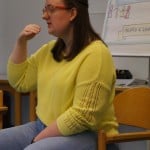 Eine Person sitzt auf einem Stuhl und gestikuliert mit ihrer rechen Hand, hinter ihr ist ein Flipchart