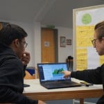 Eine junge Person zeigt einer anderen Person etwas auf einem Laptop, auf ihren Pullis steht "JuPa Emden". Im Hintergrund ist eine Moderationstafel mit vielen beschriebenen Moderationskarten.