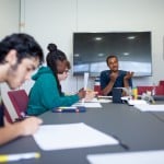 4 Personen sitzen an einem langgezogenen Gruppentisch, es liegen Zellte und Stifte auf darauf. Die Person am Kopfende ist Erwachsen und erklärt etwas, die anderen sind Jugendliche.