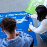 Zwei Personen knien auf dem Boden vor drei Plakaten. Sie haben Röhren in der Hand, in denen Perlen sind. Aus einer Schüssel auf einem Plakat nimmt eine Person gerade eine oder mehrere weitere Perlen. Die Plakate sind nur teilweise zu sehen, auf einem steht "Ernährung", es ist grün und eine Kuh ist im Comicstil darauf abgebildet. Auf einem blauen Plakat ist die Überschrift Langstreckenflüge und ein Flugzeug ist zu erkennen. Auf der linken Seite sind mehrere Zeilenanfänge zu erkennen, die mit "0P", "3P", "13P" und "30P " beginnen.